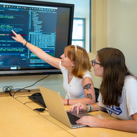 Two students study at a computer with a large TV screen displaying data