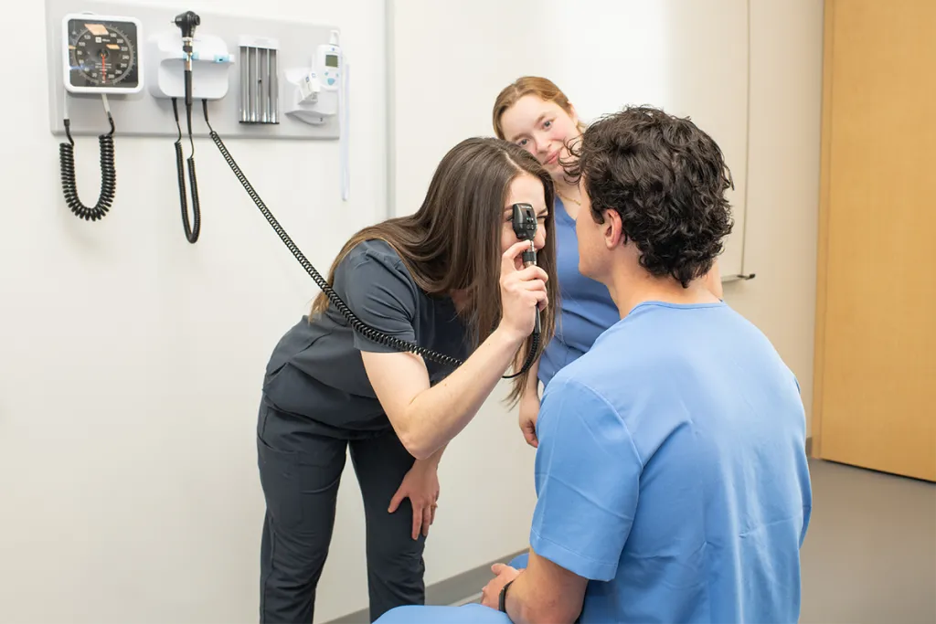 An osteopathic medicine student uses an otoscope to perform an eye exam on another student