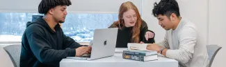 A student types on their laptop while two students review a book while sitting together at a high-top desk in front of a large window