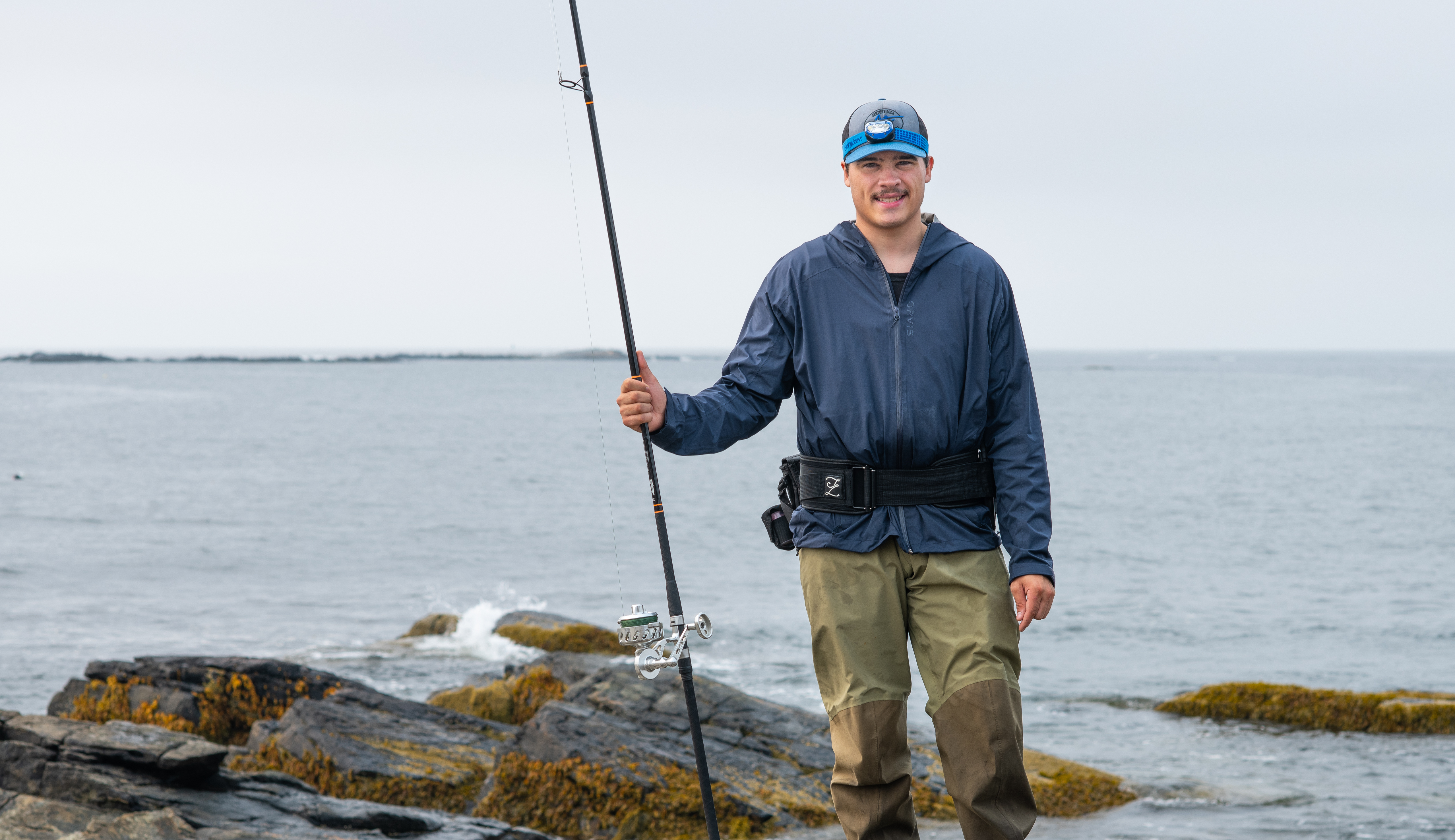 A UNE student poses with a fishing rod in front of the ocean