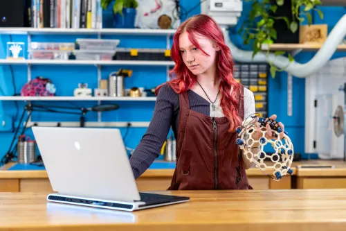 A U N E student holds up a brain wave helmet and works on their laptop