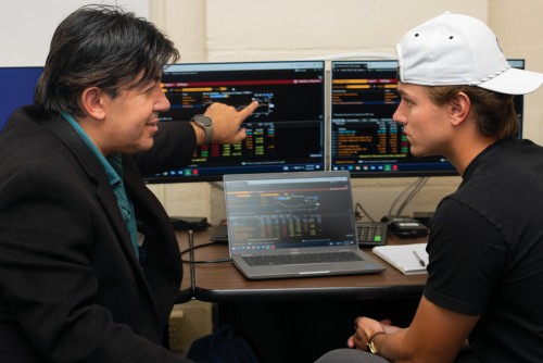 A professor and student sit in front of two computer monitors, each showing financial data