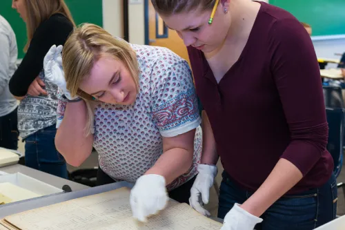 Two students wearing white gloves review cursive writing on old, yellowed paper 