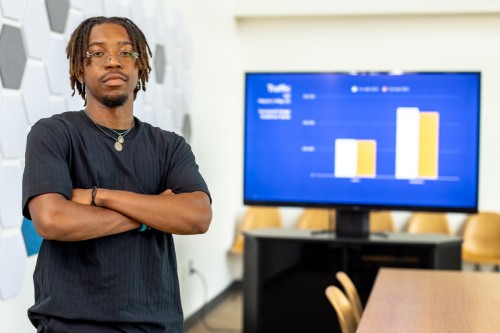 A student stands with their arms crossed in front a large computer monitor displaying a bar graph