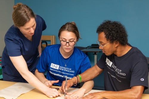 An O T student draws around a fellow student's wrist onto paper as a professor describes the process