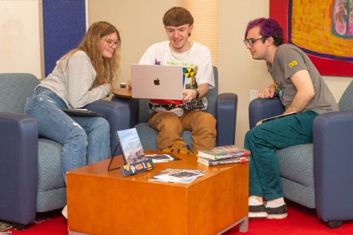 A group of three students sitting on arm chairs review material on the center student's laptop