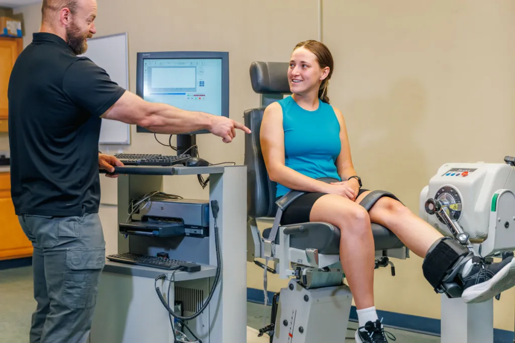 A student sits in a machine that measures muscle strength as their professor shows them the results on a monitor