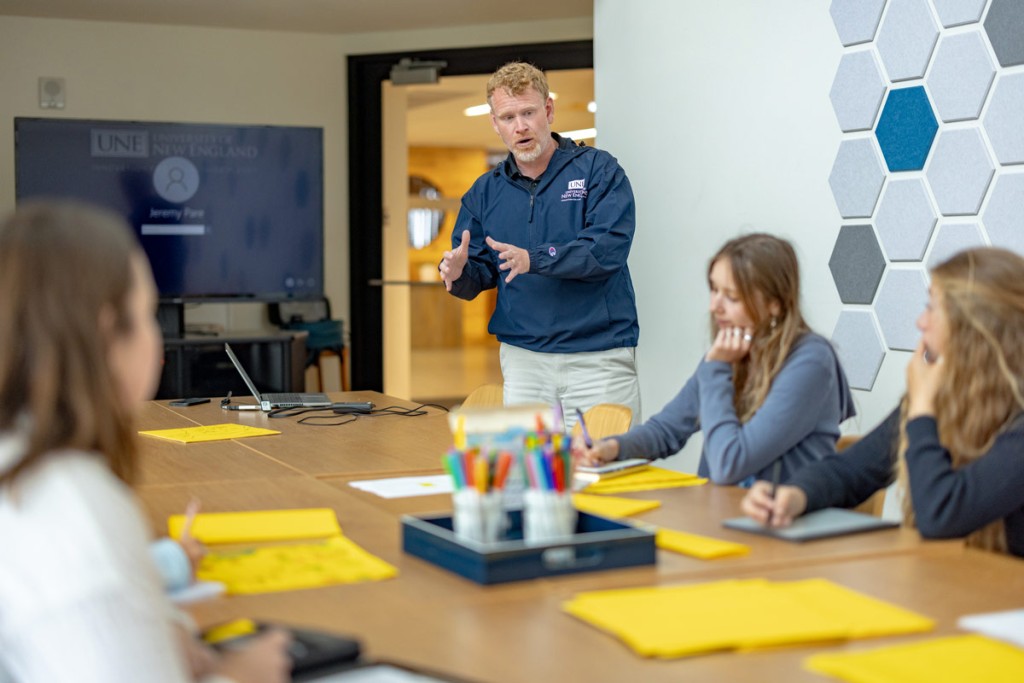 A professor speaks to a group of business students sitting at a large table with yellow paper and colorful writing utensils