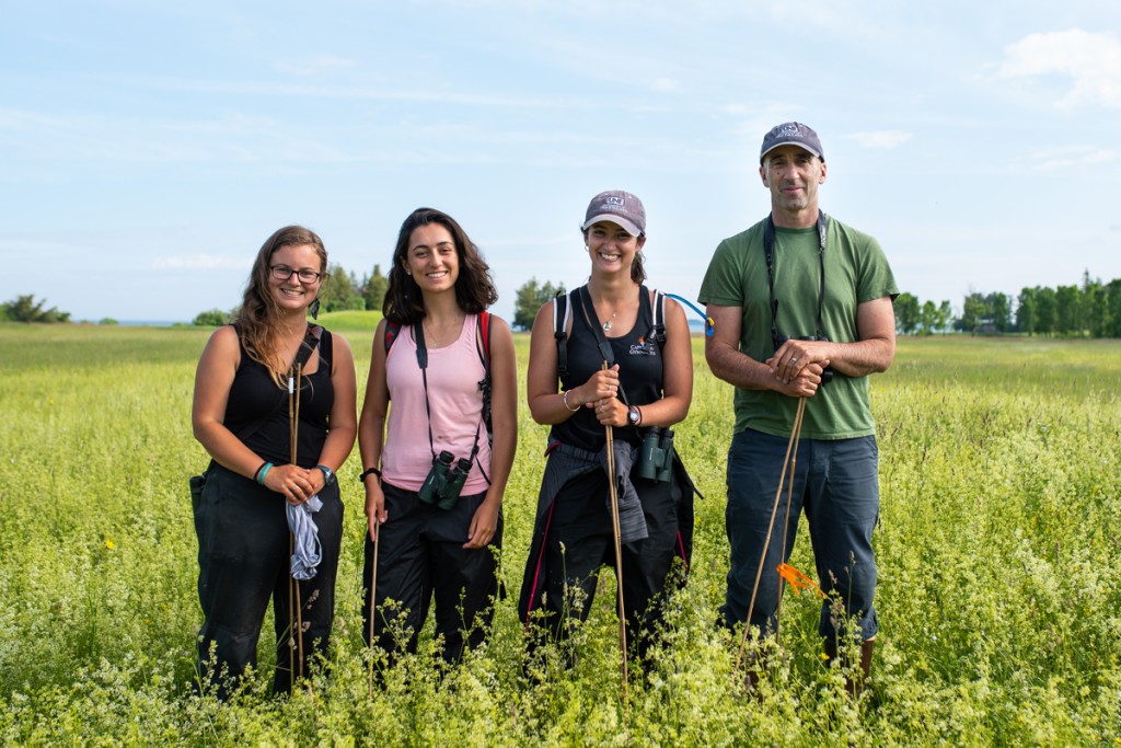 Three students wearing binoculars stand with a professor in a green field