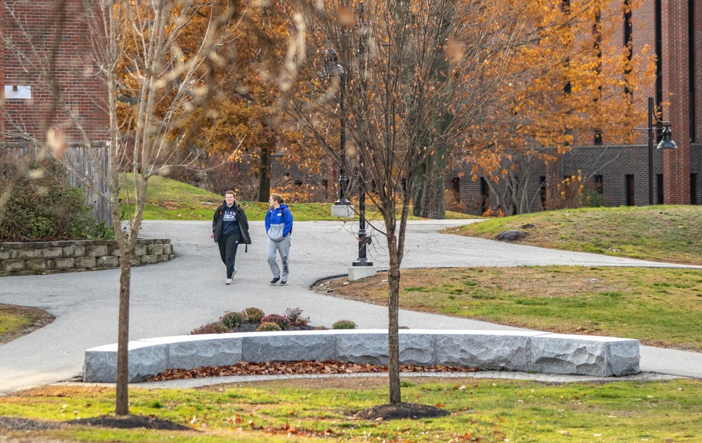 Two U N E students walk on a paved path through the greenery of Biddeford Campus