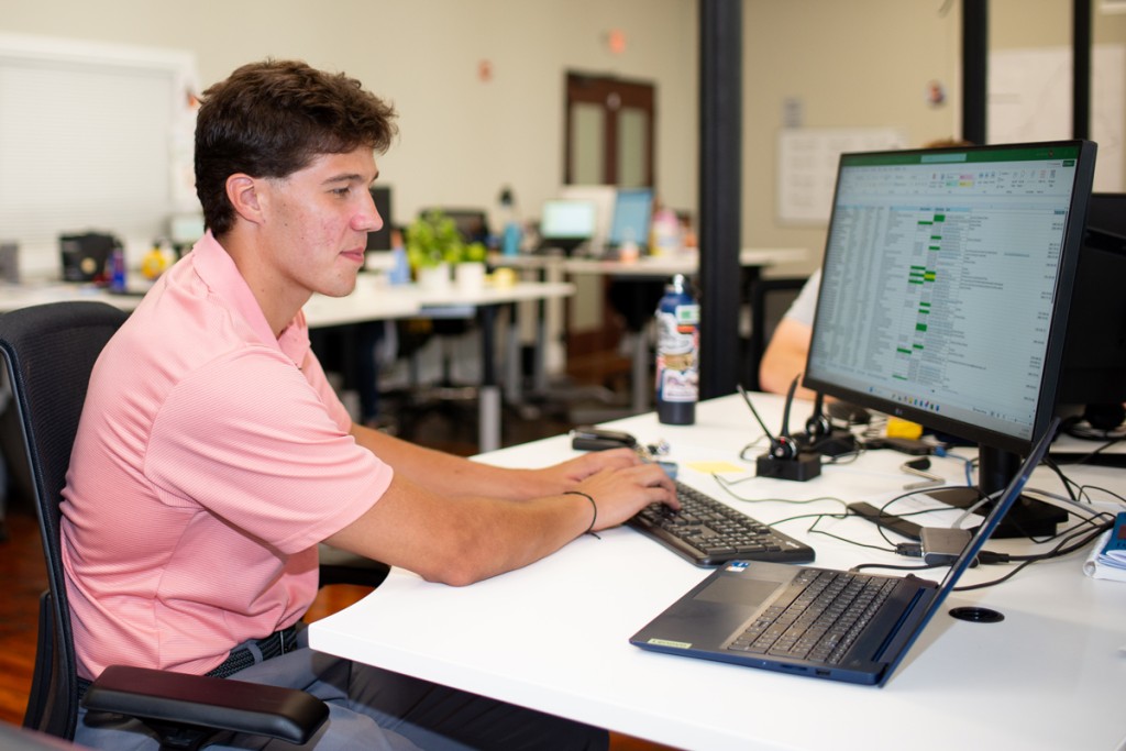 A student works at a desk with a computer displaying a spreadsheet in an office setting