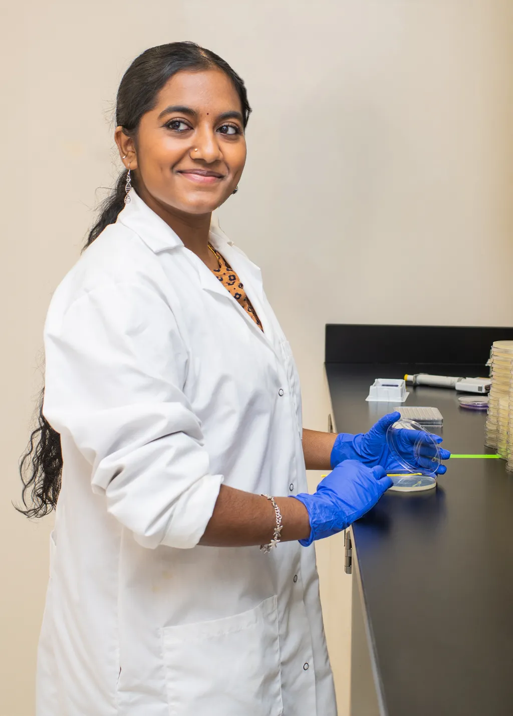 A UNE student in a white coat holds the lid to a petri dish at the counter of ther Burkholder lab while smiling toward the camera