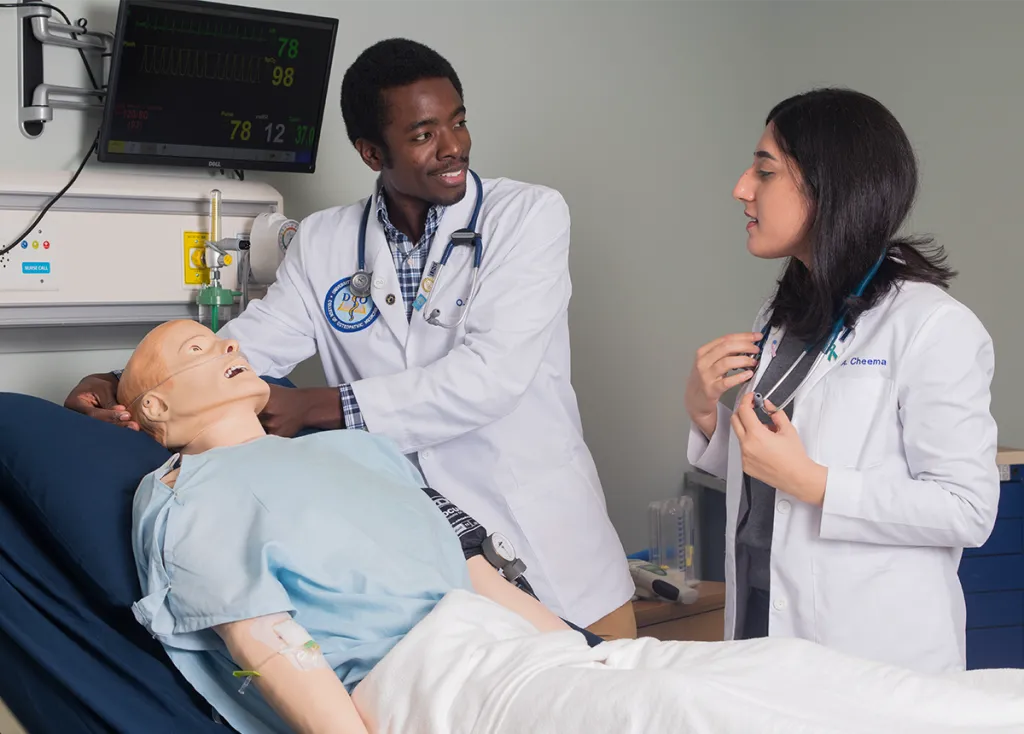 Two C O M students, in their branded white coats, work with a patient simulator