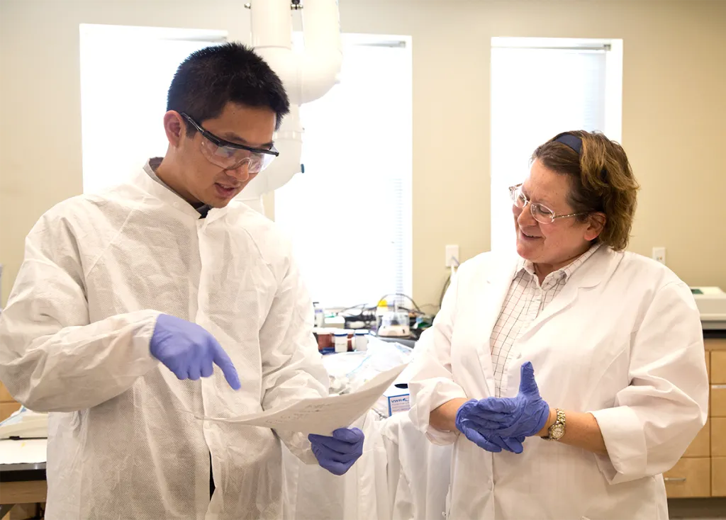 A student in a white lab coat talks to a faculty member