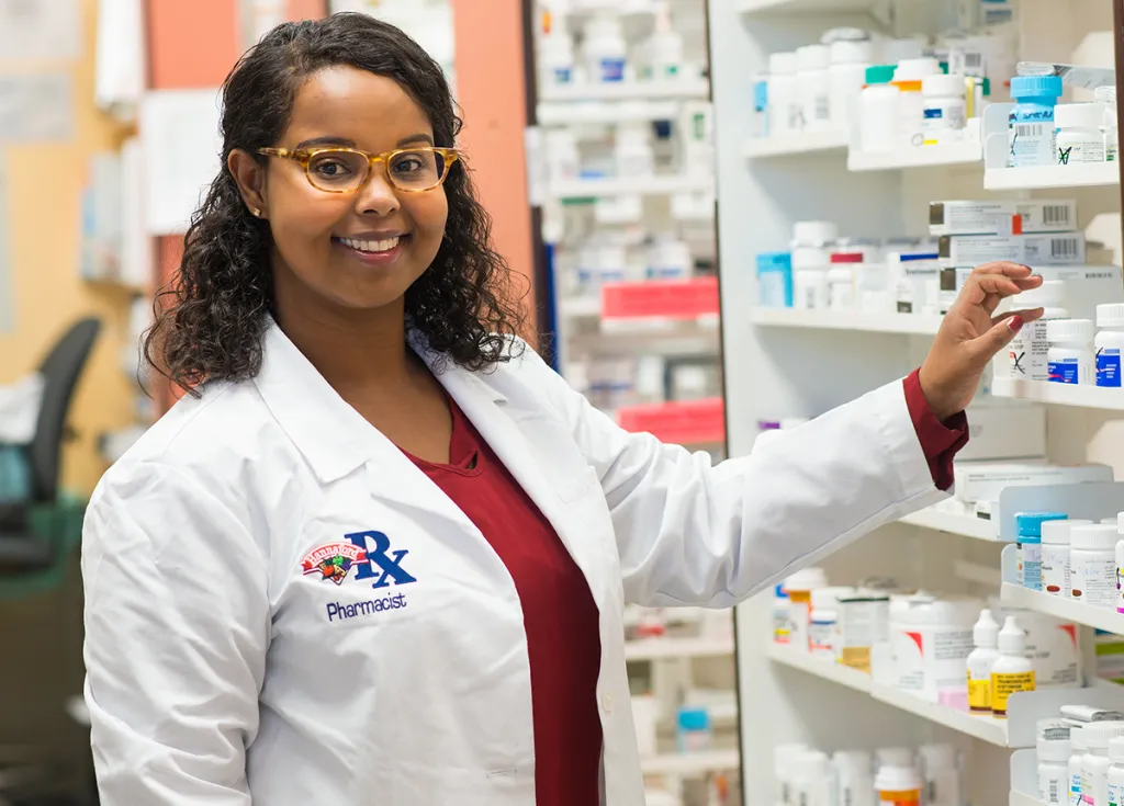 A pharmacy student smiles as she reaches for a bottle of medication from a large shelving unit