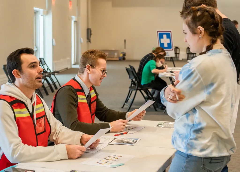 Two students in orange vests talk to a small crowd at a public health event