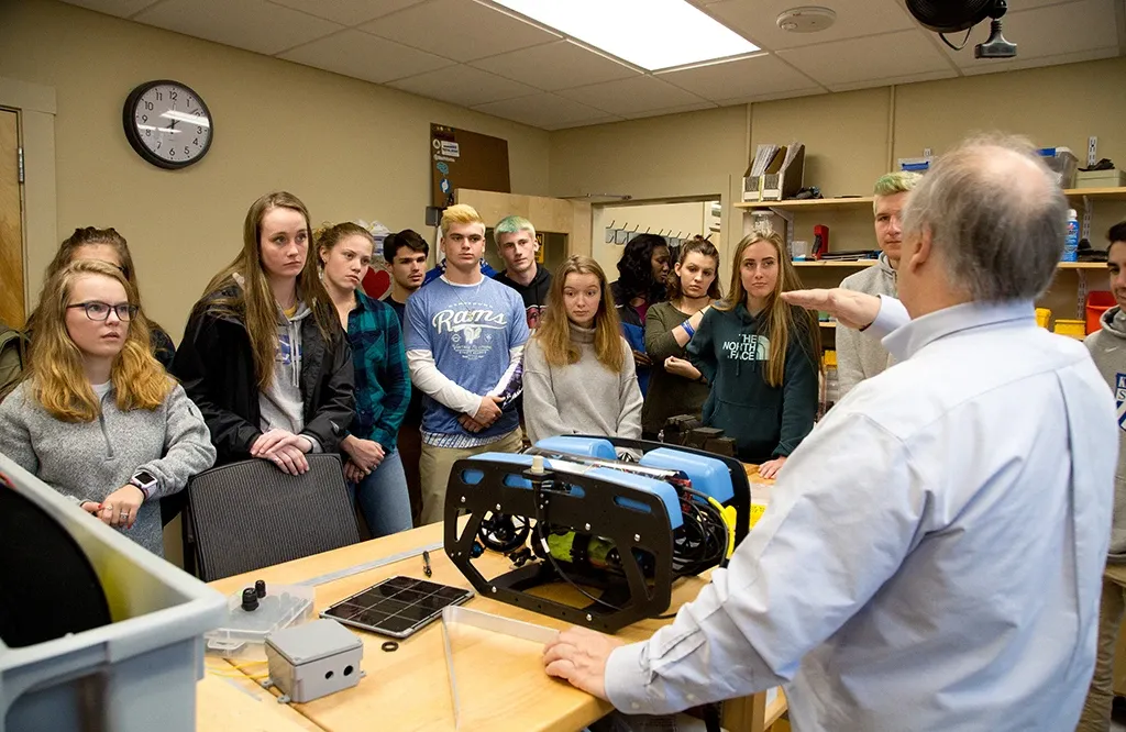 A group of high school students gathered around a professor as he explains a marine science tool produced by U N E students