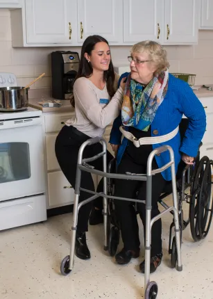 A student assists an elderly person move from their wheelchair to their walker