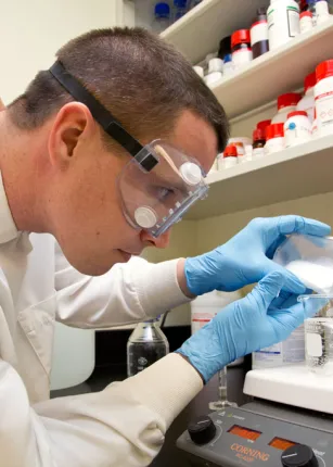 A C O M student pours a substance into a liquid-filled beaker on a scale