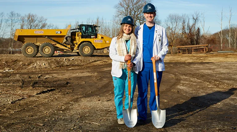 Two UNE COM students pose with shovels, the construction site in the background