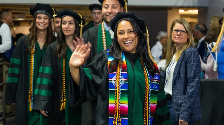 A UNE graduate wearing doctoral regalia waves walking into the ceremony