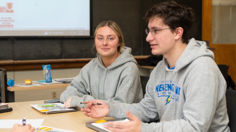 Two students discuss a book in a class with a blackboard behind them