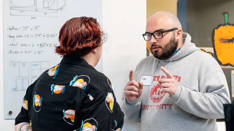 Two students from the innovation team discuss measurements of a project written out on a whiteboard