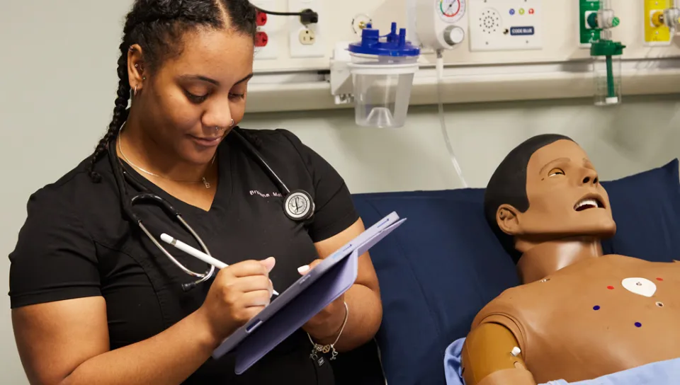 A physician assistant student wears a stethoscope and takes notes on a tablet while in a sim lab class