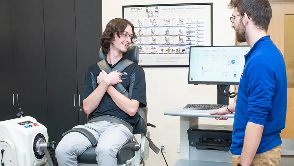 One athletic training student sits upright in a machine that tests leg strength while another student looks at the results on a computer monitor