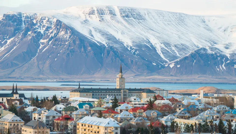 A town with colorful buildings is set before a snowy mountain range under a cloudy sky