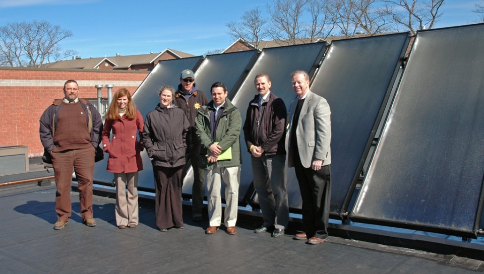 A group of U N E faculty and staff stand in front of five large solar panels