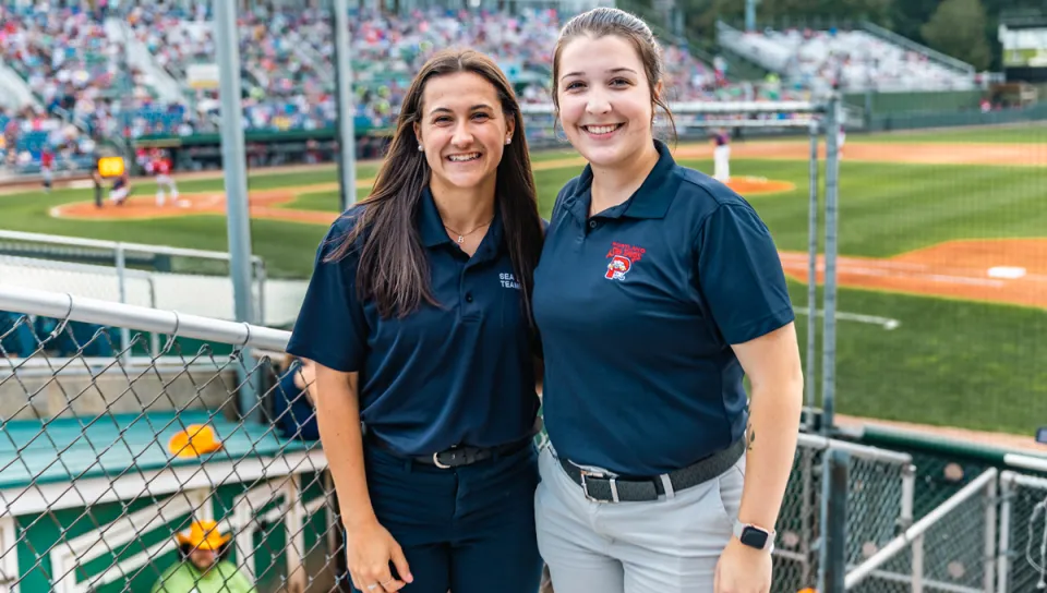 Two students wearing Sea Dogs polo navy shirts stand in front of Hadlock Field