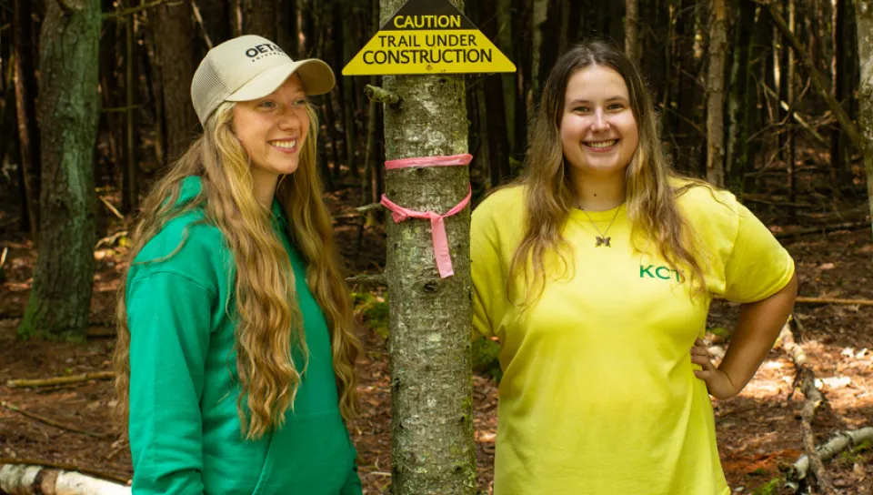 Two students standing in the woods next to a sign that reads, "Caution: Trail Under Construction"