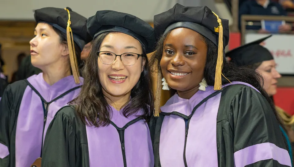 Two U N E graduate students smiling for the camera while other graduates are visible in the background