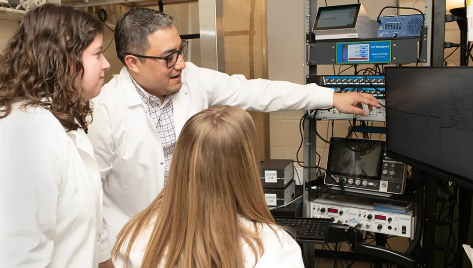 Queme in a white coat points at a screen and expains data to two UNE students who are also in a white coat