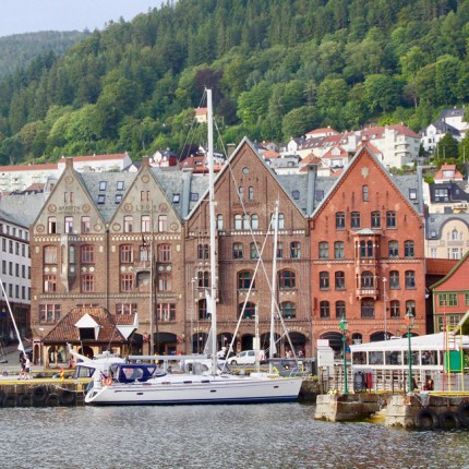 A boat drives through a river with brick buildings and mountains in the background