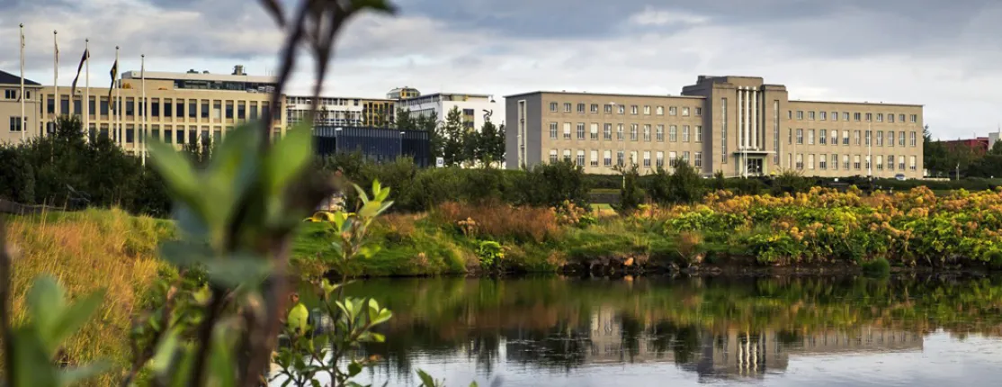 A scenic view of a large building with a pond in the foreground and greenery around, set against a cloudy sky