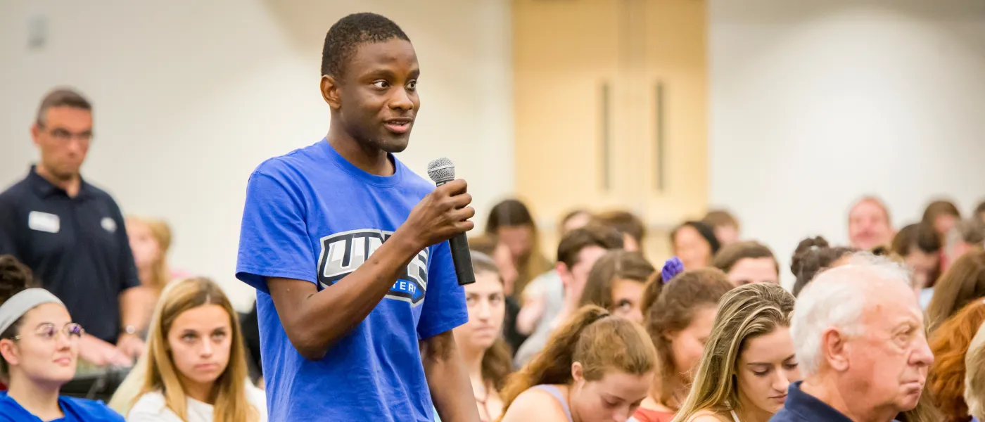 A student in a blue U N E t-shirt asks a question from the crowd of a President's Forum event