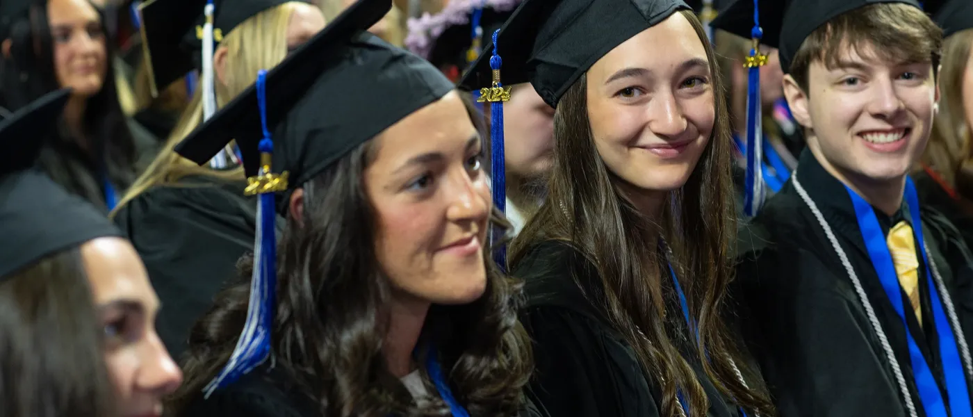 Two U N E graduate students look at the camera in a the crown of graduates before they listen to commencement speaker Bernie Sanders