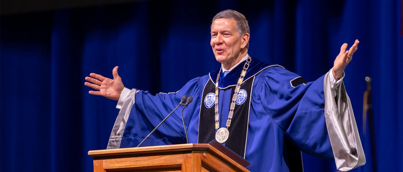 U N E president James D. Herbert stands at podium at graduation in the commencement garb with his arms outstretched gesturing to the crowd