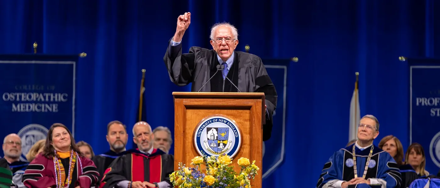 Bernie Sanders standing at the podium raises a hand gesturing to the crowd at the UNE 2024 commencement ceremony