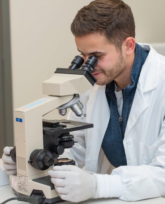 A student in a white lab coat peers into a microscope