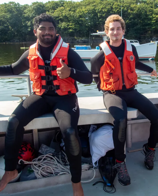 Two marine science students wearing wetsuits and orange life vests sit on the edge of a boat