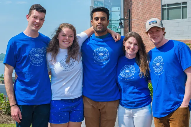 Five U N E students wearing matching blue U N E t-shirts pose happily