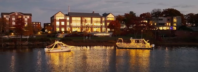 Two boats covered in white holiday lights sit in the water in front of the Ripich Commons on U N E's Biddeford campus