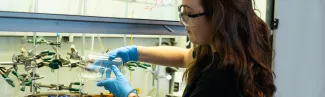 A student wearing safety glasses in a lab pours liquid from a beaker into a petri dish 