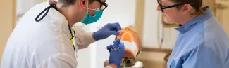 Two dental hygiene students practice injecting medicine into the gums of a patient simulator