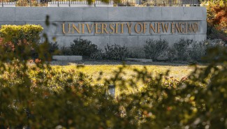 A stone sign bearing the name "University of New England" is seen through the trees