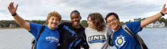 A group of four students in U N E t-shirts pose in front of the ocean off of the Biddeford Campus