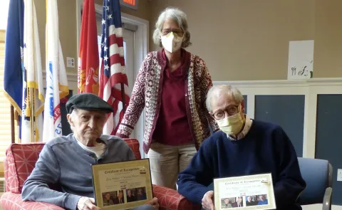 Photo of two veterans sitting in chairs with awards and Susan Gold posing with them